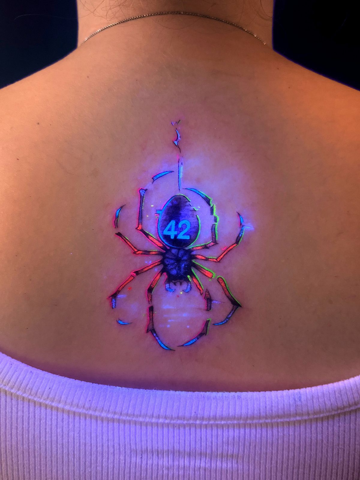 Neon glowing spider tattoo on a person's back under UV blacklight.