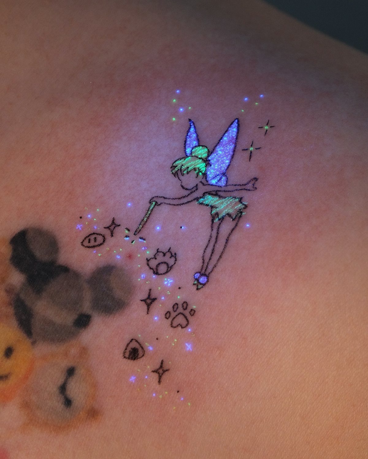 Tinker Bell fairy tattoo glowing in iridescent UV ink on a person's skin.