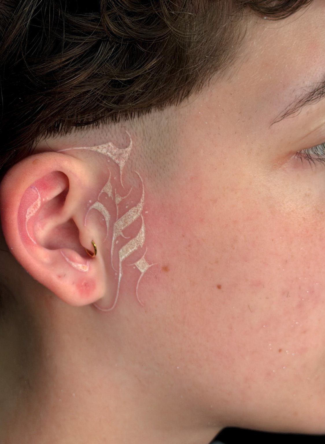 White ink tattoo with ornamental swirls on the skin beside a person's ear.