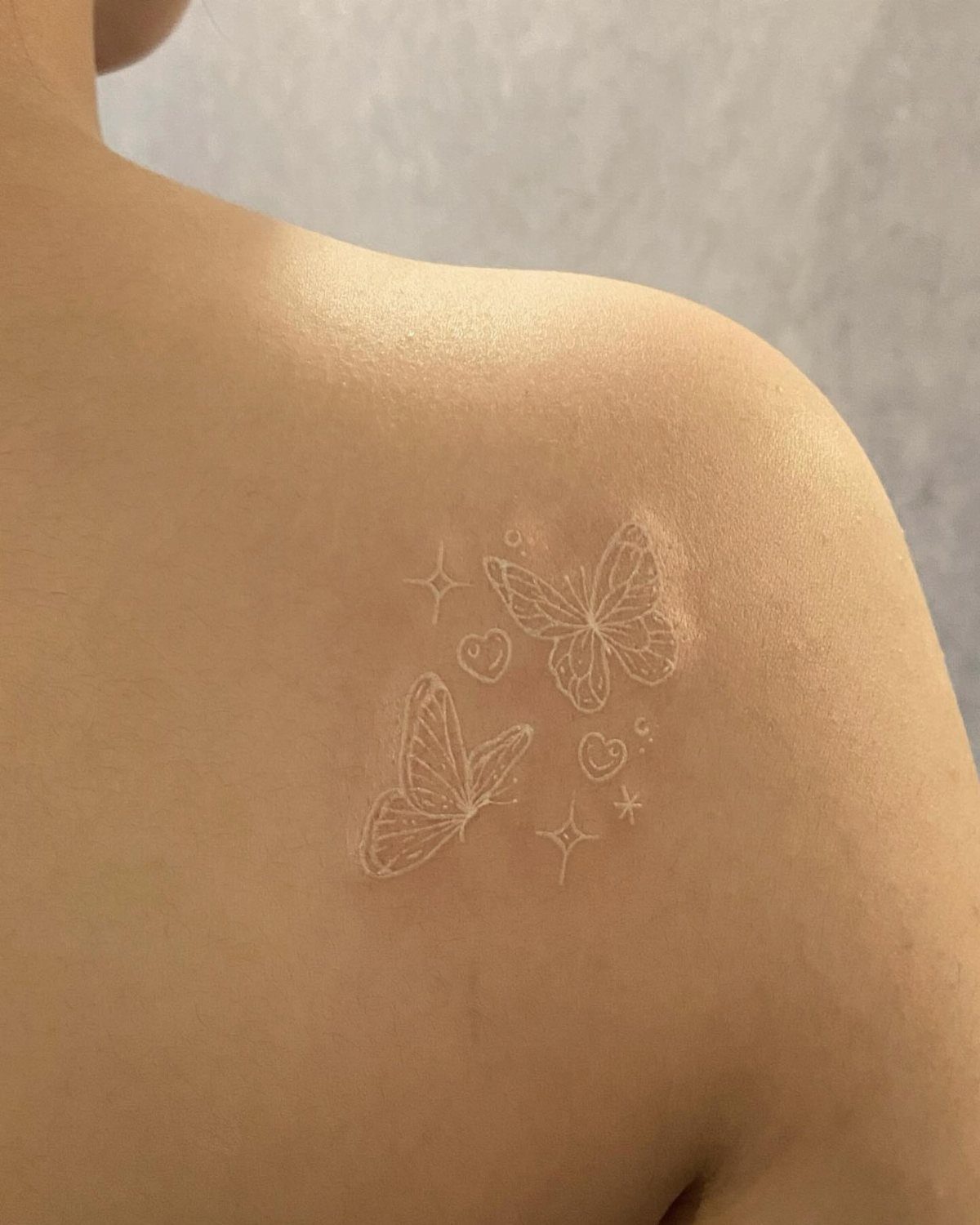 White ink tattoo of two butterflies and stars on a person's shoulder.