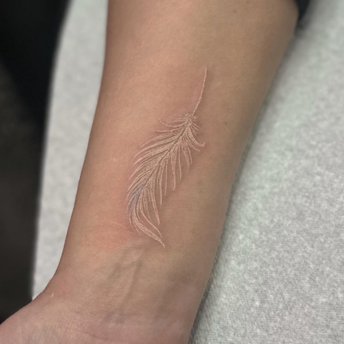 White ink tattoo of a detailed feather on a person's inner forearm.