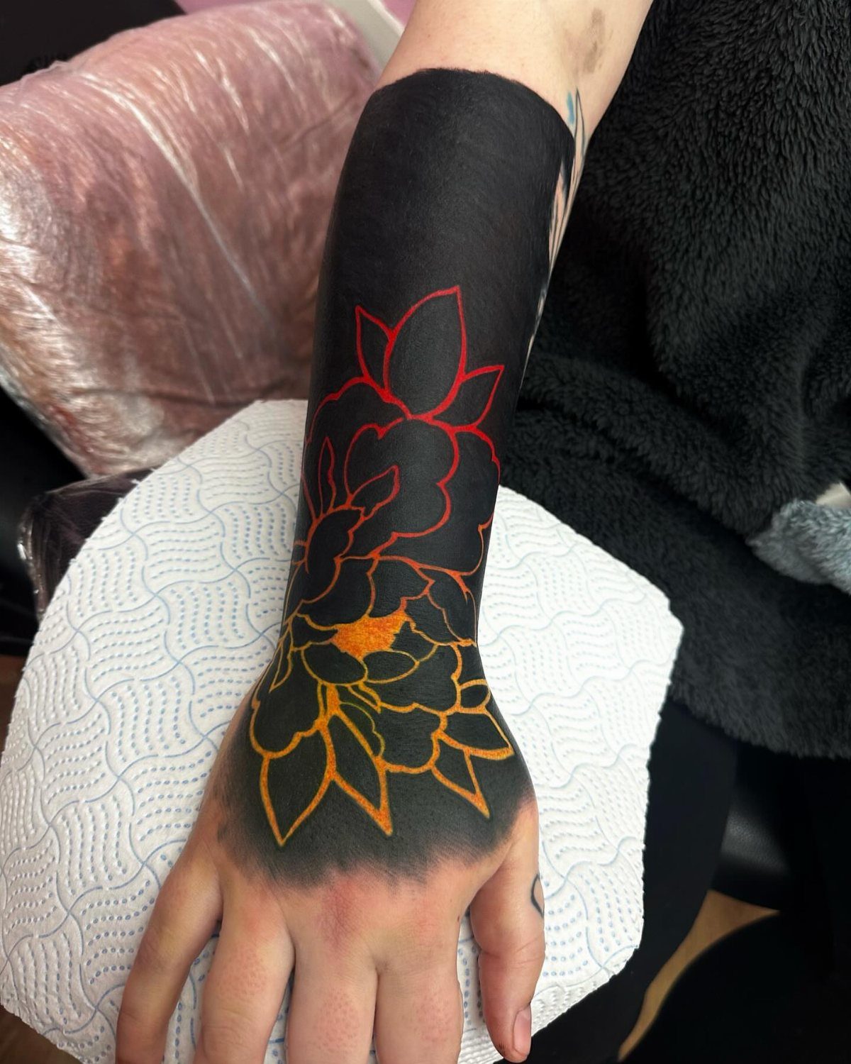Blackout tattoo on a forearm with orange floral outlines layered over the black ink.